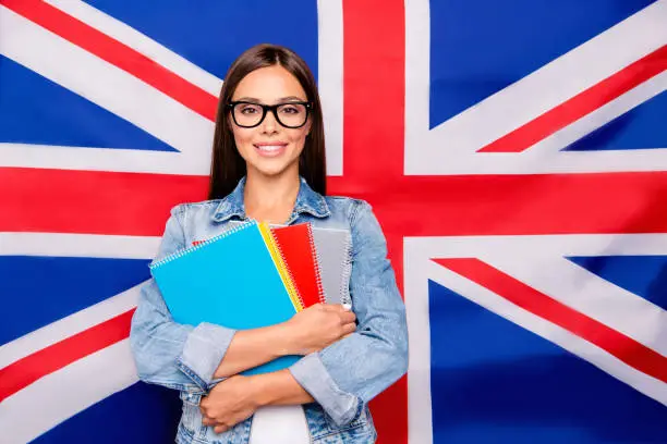Portrait of cute sweet lovely confident smiling student lady emigrant wearing spectacles, casual denim jeans, holding notepads, isolated over Great Britain union jack flag