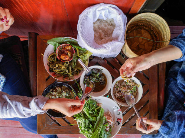 Two Thai women traditionally eating a selection of freshly cooked Northern Thai food of fresh vegetables, soups and curries served in dishes on a wooden table, along with sticky rice, which is a food staple in Northern and Northeast Thailand. dos mujeres tailandesas tradicionalmente comer una selección de recién norte tailandés el alimento cocinan de verduras, sopas y curry servido en platos de una mesa de madera, junto con el arroz pegajoso, que es un alimento básico en el norte y noreste