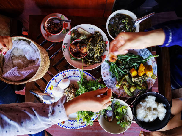 two thai women traditionally eating a selection of freshly cooked northern thai food of vegetables, soups and curries served in dishes on a wooden table, along with sticky rice, which is a food staple in northern and northeast thailand. - bamboo shoot imagens e fotografias de stock