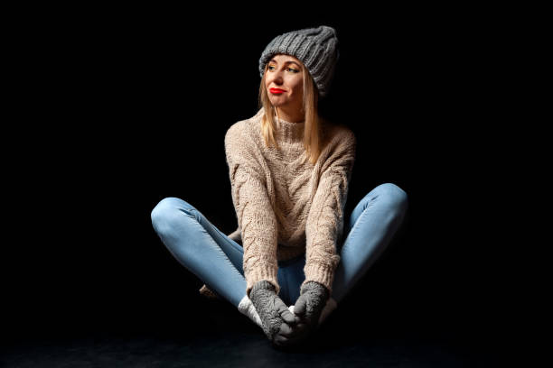 giovane bella ragazza bionda in guanti a maglia e cappello in grigio, jeans blu, maglione beige si siede sul pavimento in una posa farfalla baddha konasana e distosta lo scenografo su uno sfondo nero isolato - butterfly women humor fun foto e immagini stock