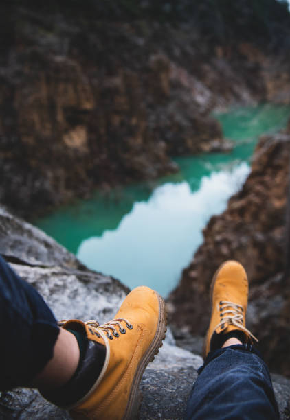 Feet of traveler or wild nature explorer in yellow dirty hiking boots sitting over chasm with green mountain river canyon at background. Travel, tourism, wanderlust or adventure concept