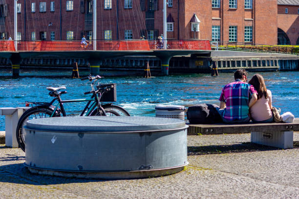 The quayside near the Black Diamond National library in Copenhagen, Denmark with the quayside in the foreground and a young couple enjoying the late afternoon spring sunshine at the side of the canal at sunset.