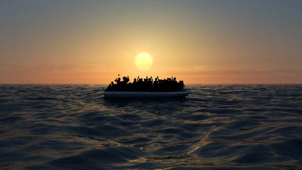 A crowded boat carrying people drifts on the open sea at sunset, silhouetted against the golden sky.