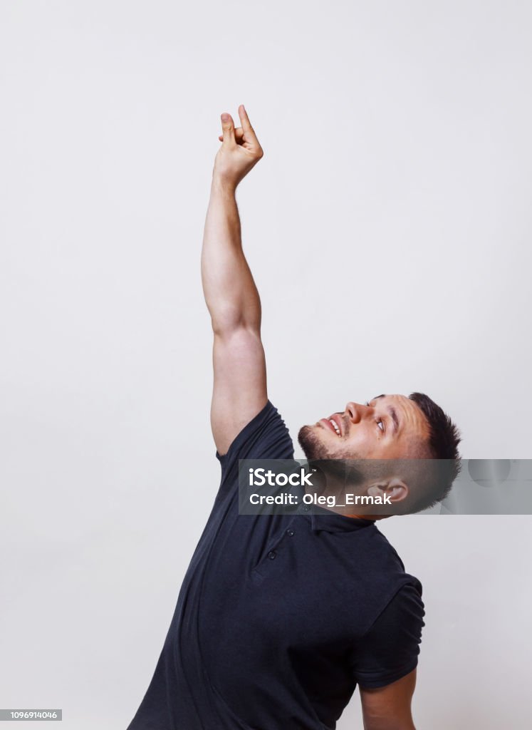 Grab gesture or climb something. Portrait of casual man wearing a darck shirt stretches his hand up ase wants to get something or he wants to screw in a light bulb on grey background High Up Stock Photo
