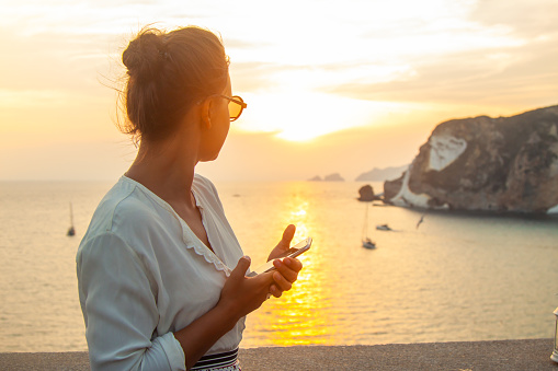 Young woman with smartphone looking at sunset in front of the sea on Ponza island coast, on a wall with view of the ocean. 9871