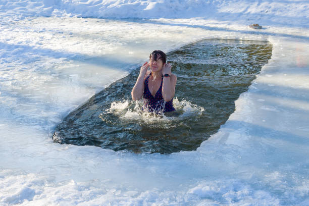 A woman emerges from the ice hole. Baptism day The holiday of Baptism is celebrated by all Orthodox Christians on January 19. On this day, there is a tradition to dip three times into a hole with ice-cold water. Baptism is the first and most important Christian sacrament. It is recognized by all Christian denominations, although not in the same sense. Through baptism, a person becomes a member of the Church. Only after baptism is a person given the opportunity to participate in all the other sacraments of the Church, and above all, in the Eucharist. The sacrament of baptism consists either in immersion (single or triple - depending on the confession) of a person in water, or in dousing a person who receives baptism, with the prayer pronounced by the priest. woman cold water swimming stock pictures, royalty-free photos & images