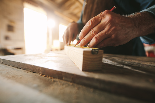 Close up of hands of senior carpenter doing some markings on wooden bar with measuring tape and pencil.