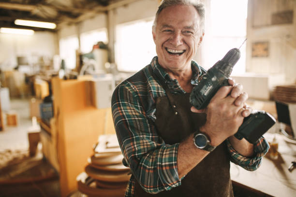 Smiling senior carpenter holding drill machine Smiling senior carpenter holding drill machine against workshop. Mature male worker smiling confidently to the camera holding drilling machine at his carpentry workshop. carpenter portrait stock pictures, royalty-free photos & images