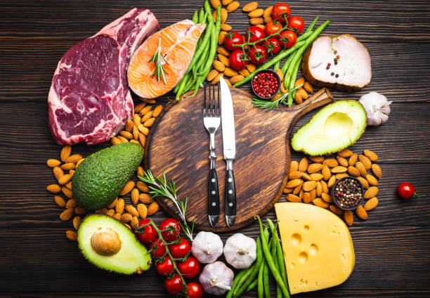Keto diet foods Knife and fork over wooden cutting board and ketogenic low carbs ingredients for healthy eating concept and weight loss, top view. Keto foods: meat, fish, avocado, cheese, vegetables, nuts ketogenic diet stock pictures, royalty-free photos & images