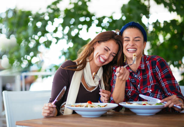 Two female friends eating salad and laughing Photo of beautiful women eating fresh salad at restaurant brunch photos stock pictures, royalty-free photos & images