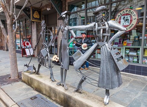 Asheville, NC, USA-1/18/19: The Shopping Daze metal sculpture standing in front of Malaprops Bookstore, with a banjo-playing busker visible behind The artwork is by sculptors Tekla and Dan Howachyn and Tucker Cooke, and was installed to represent the downtown area of the 1970s.