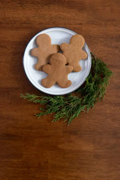 Three gingerbread men arranged on a white plate with evergreen surronding it on a cherry table