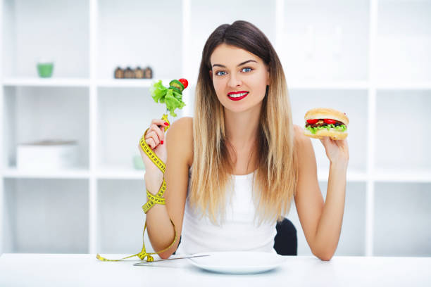 un régime concept, belle jeune femme, choisir entre une alimentation saine et la malbouffe - food measuring hamburger dieting photos et images de collection