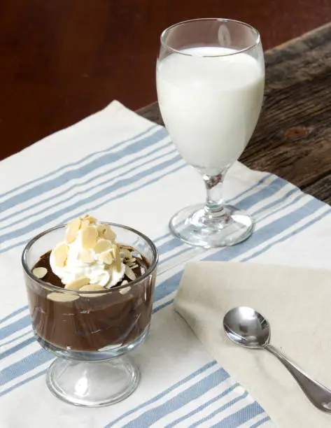 a cup of chocolate pudding with whipped cream on top and almond slices arranged on a placemat on a wood table with a spoon and glass of white milk