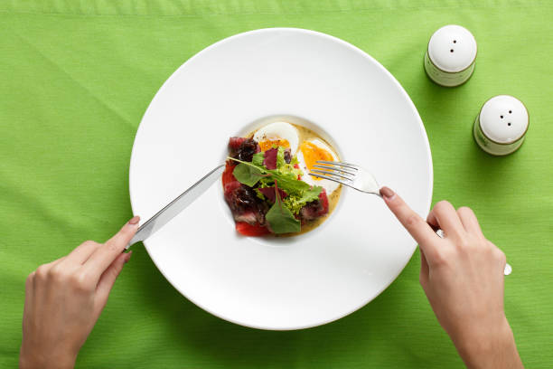 Top view of dish eggs with meet and salad on plate. Woman hands holding fork and knife. Dish on white plate on green background, view from above. Restaurant dish, healthy food concept