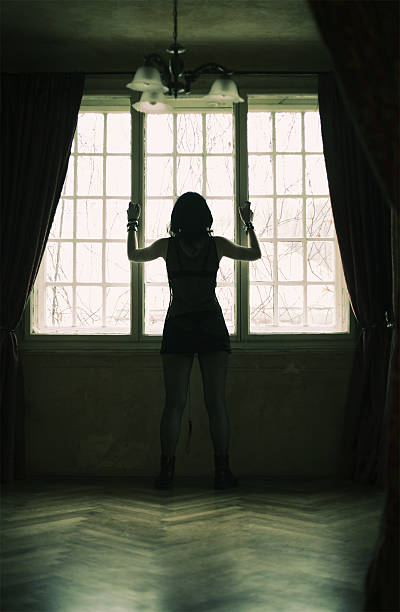 A lonely young girl looking thru the window in old, abandoned castle room.