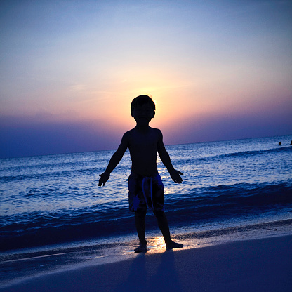 Panoramic silhouette shot of an Indian kid looking at the orange sky during the sunset. Hopeful kid stares at the sun. Kid lost in deep thoughts after looking at the sunset.