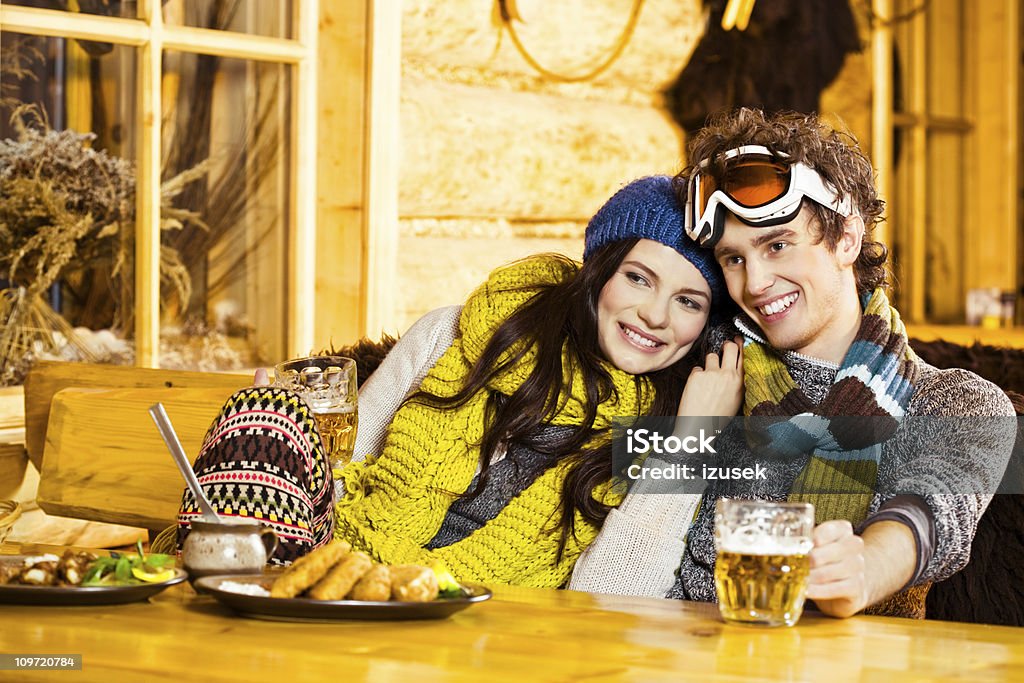 After skiing A cute young couple wearing a winter clothes resting after skiing. They sitting in the mountain restaurant, eating and drinking beer. 20-24 Years Stock Photo