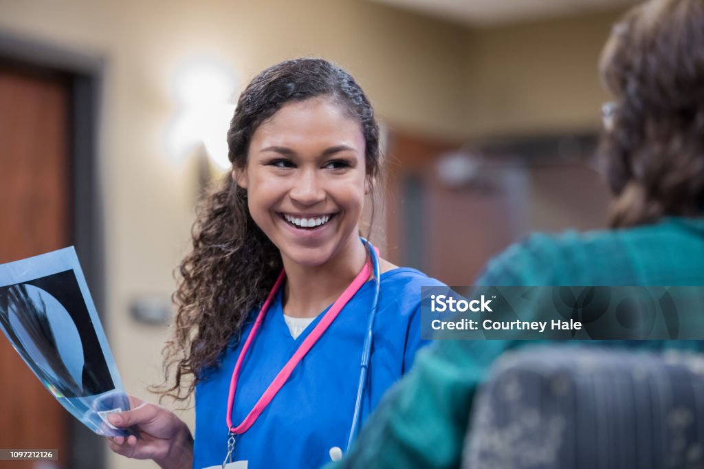Radiologist or nurse smiles while discussing x-ray with patient in clinic Young adult mixed race woman smiles while holding an x-ray and discussing it with a patient in radiology clinic or hospital. Radiologist Stock Photo