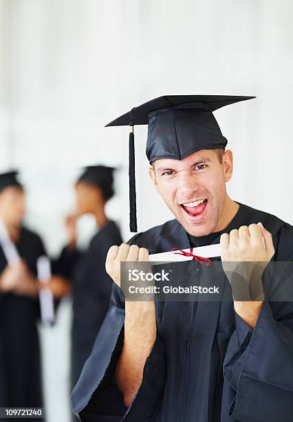 Excitación Hombre Que Agarra Graduación Certificado Con Amigos En El Fondo Foto de stock y más banco de imágenes de 20-24 años