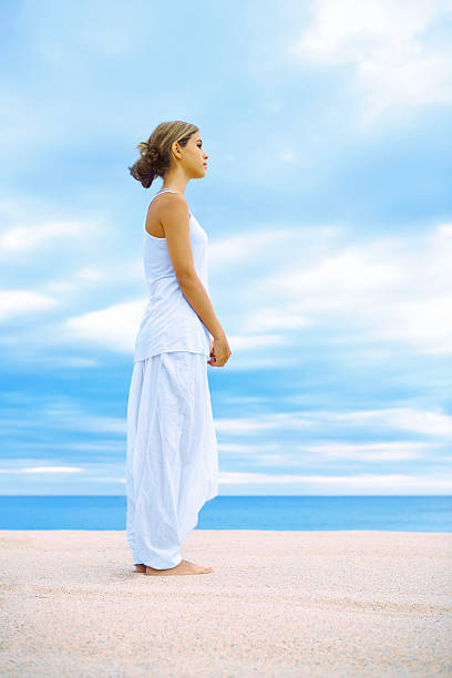 A woman is standing on the beach with her feet in the water and holding a drink. The scene is peaceful and relaxing, with the sound of the waves and the ocean breeze