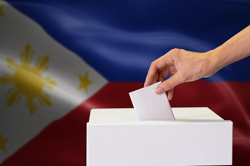 Close-up of human hand casting and inserting a vote and choosing and making a decision what he wants in polling box with Philippines flag blended in background.