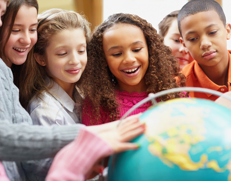Woman in glasses and positive children examining globe while standing around table in geography classroom during lesson at school
