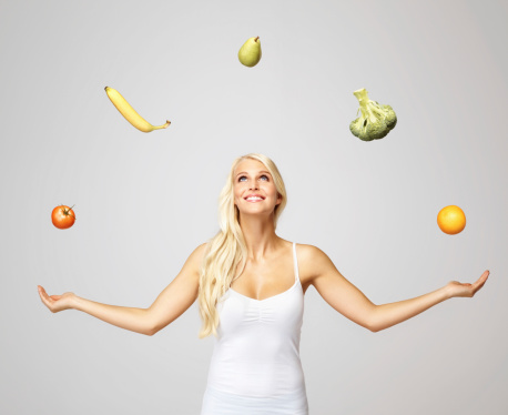 Portrait of a smiling pretty woman juggling fruits and vegetables against grey background