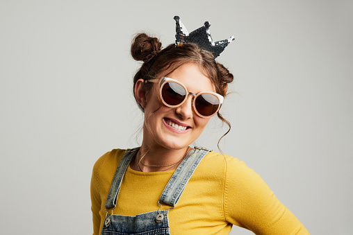 Studio shot of an attractive young woman wearing sunglasses and a crown against a grey background