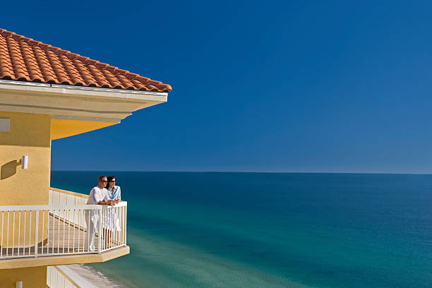 Couple Standing on Balcony Looking At The Ocean Young man and woman standing on balcony looking out across the ocean. pretty beach house stock pictures, royalty-free photos & images