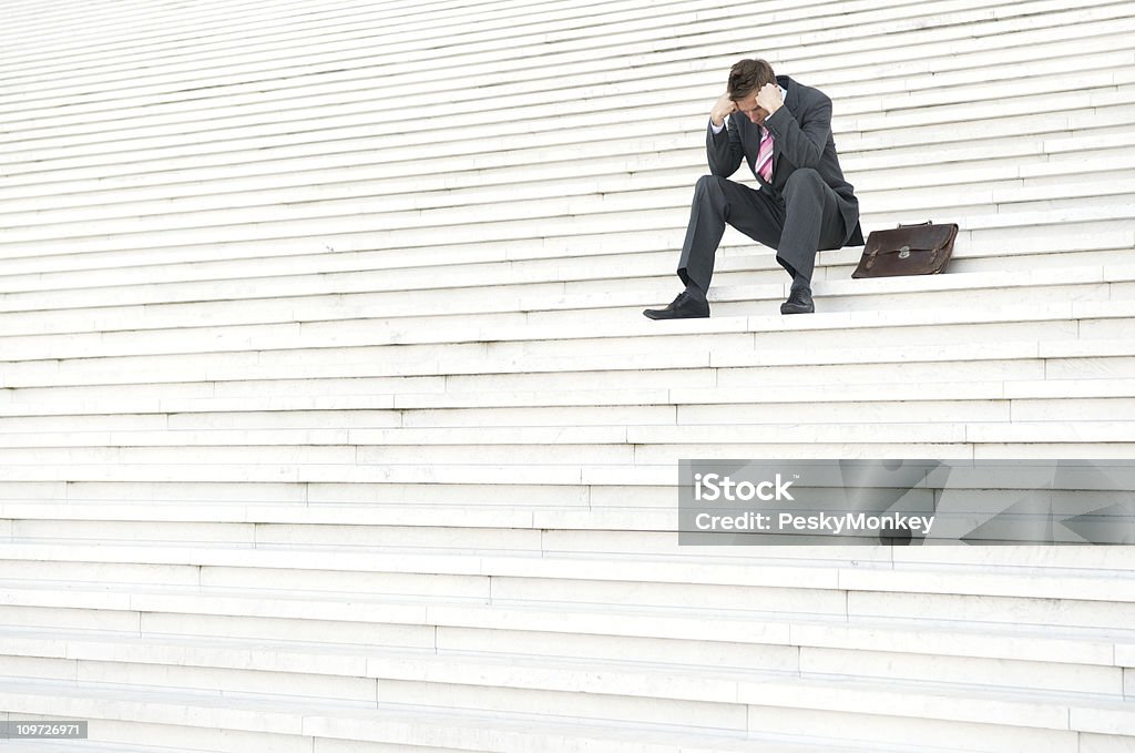 Depressed Businessman Sitting Outdoors on Steps with Head Down Depressed businessman looks small outdoors on a big white staircase, looking down with his briefcase by his side Adversity Stock Photo