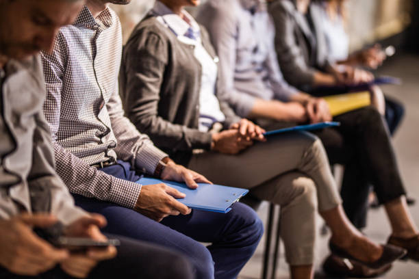 Group of unrecognizable candidates waiting for a job interview. Large group of unrecognizable business people waiting for a job interview. Focus is on man with blue clipboard. interview event stock pictures, royalty-free photos & images