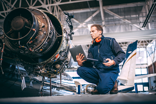 Aircraft engineer in a hangar holding a laptop computer while repairing and maintaining an airplane jet engine.