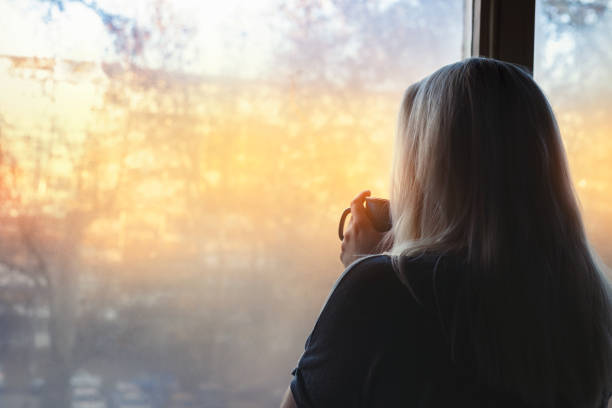 mujer rubia de pie junto a la ventana, con la taza de café en las manos, mirando a la luz de la mañana - women depression window sadness fotografías e imágenes de stock