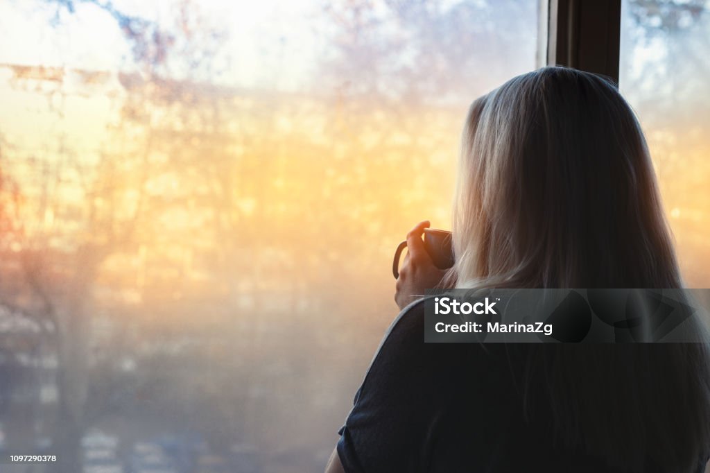 Blonde woman standing by the window, with coffee cup in hands, looking out into the morning light One Woman Only Stock Photo