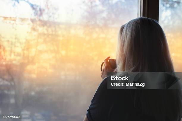 Blonde Woman Standing By The Window With Coffee Cup In Hands Looking Out Into The Morning Light Stock Photo - Download Image Now