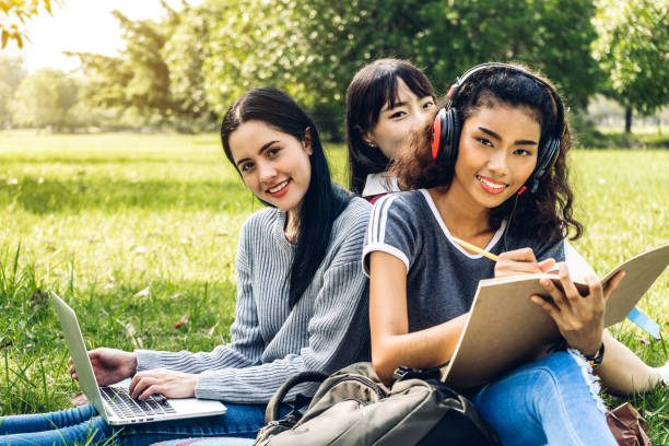 istock Group of smiling international students or teenagers sitting and using laptop computer doing their homework with book together in park at university.Education and friendship Concept 1097296524