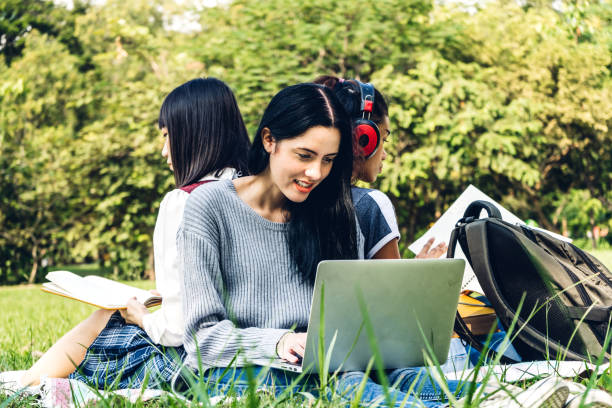 istock Group of smiling international students or teenagers sitting and using laptop computer doing their homework with book together in park at university.Education and friendship Concept 1097296530