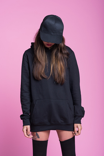 Studio portrait of worried young woman wearing black hooded shirt, knee socks and baseball cap, standing against pink background.