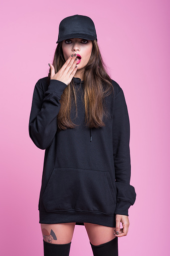 Studio portrait of beautiful young woman wearing black hooded shirt, knee socks and baseball cap, standing surprised against pink background.