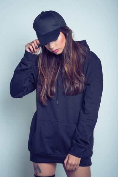 Sad young woman in black clothes against grey background Studio portrait of worried young woman wearing black hooded shirt, knee socks and baseball cap, standing against grey background. woman wearing baseball cap stock pictures, royalty-free photos & images
