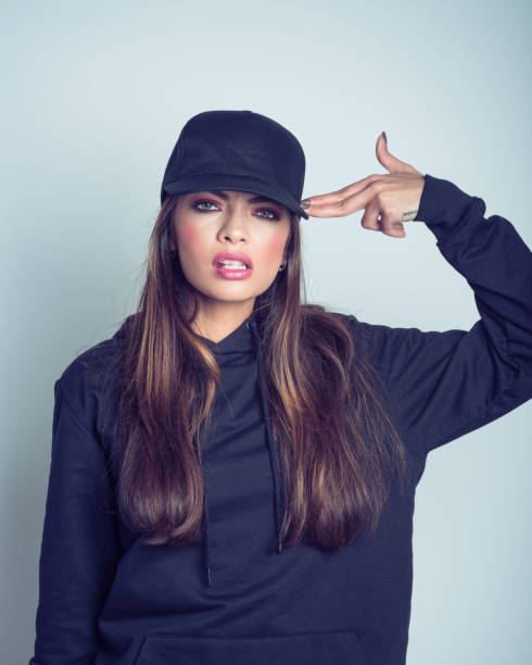 Studio portrait of beautiful young woman wearing black hooded shirt and baseball cap, standing against grey background and making suicide gesture.