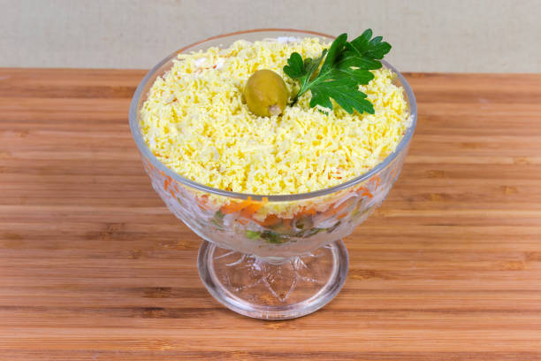 Fish salad with cheese, boiled eggs and vegetables, sprinkled with crumbled egg yolk and decorated with olive and parsley, known in post-Soviet states cuisines as mimosa salad, in glass salad bowl