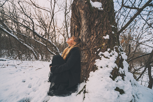 Lonely woman leaning on tree in forest in winter.