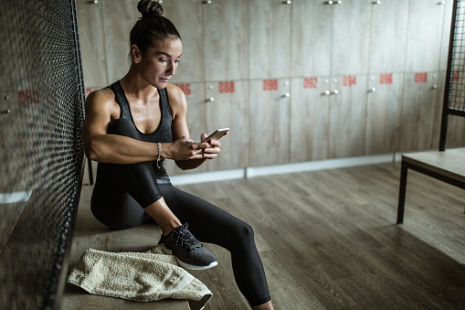 Young athletic woman using mobile phone after sports training at gym's dressing room.