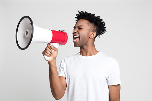 Portrait of afro american man yelling into a megaphone on gray background. African male making an announcement with megaphone.