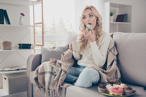 Portrait of nice lovely attractive funny hungry wavy-haired lady holding in hands cup eating large plate of tempting seductive sweets enjoying life in light interior room