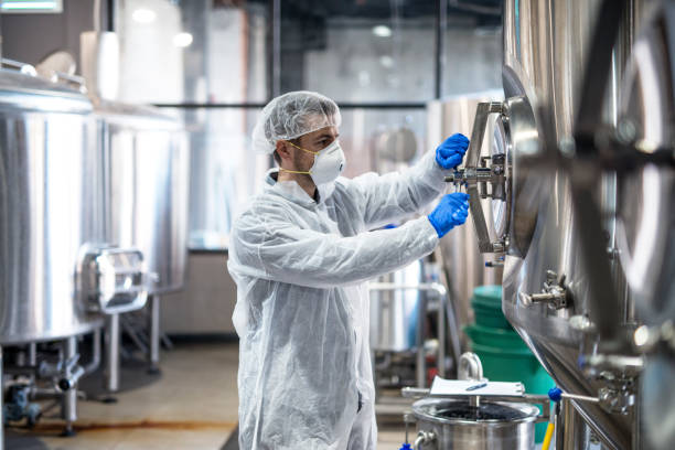 istock Technologist in protective uniform wearing hairnet mask goggles and gloves closing industrial tank reservoir. 1097339540