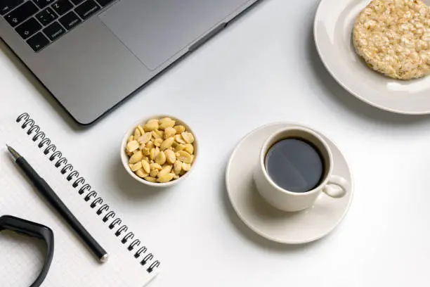 Healthy snacking at work during break time. Crispy rice rounds with peanuts, cup of coffee near the laptop, fitness-tracker and notebook. White organized desk.