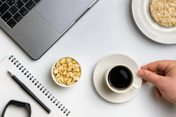 Healthy snacking at work during break time. Sportsman eating crispy rice rounds with peanuts, cup of coffee near the laptop, fitness-tracker and notebook. White organized desk.
